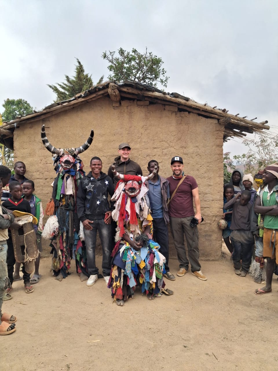 Guests posing with horned Gule Wamkulu dancers outside a mud-brick house